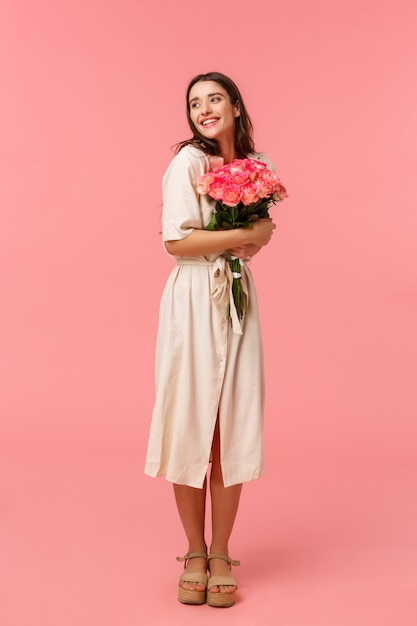 Full-length vertical portrait romantic ecstatic young woman receiving touching gift, embracing cute bouquet roses, looking dreamy and delighted, smiling thoughtful, pink wall