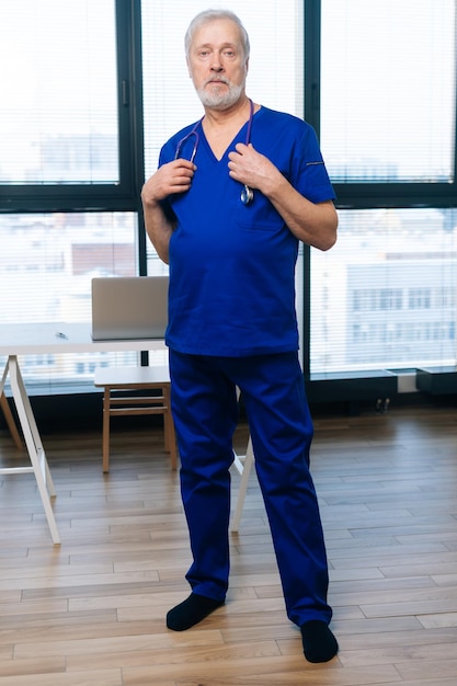 Full length vertical portrait of serious senior adult male doctor in medical uniform standing in hospital office on background of window looking at camera