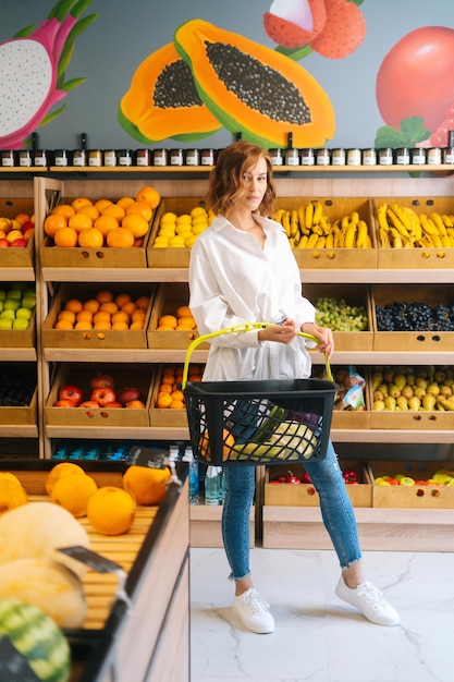 Full length vertical portrait of young woman customer holding basket of fruits standing at fruit and