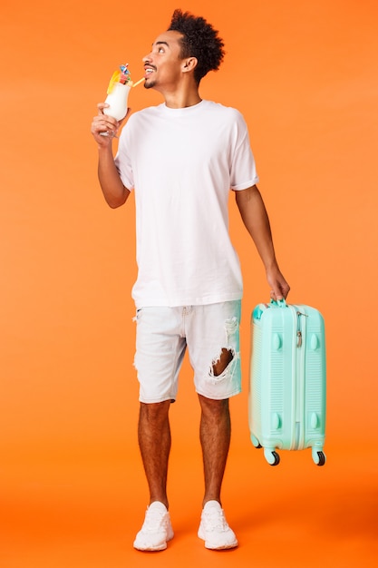 Full-length vertical shot dreamy happy and relaxed african-american man carrying luggage, suitcase and drinking cocktail, looking around pleased and smiling, book in luxury hotel, orange wall