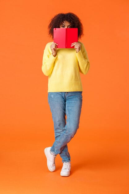 Full-length verticale opname verlegen en schattig Afro-Amerikaanse moderne stedelijke meisje met afro kapsel, gezicht verbergen achter rode laptop en gluren camera, schrijven in dagboek, staande oranje