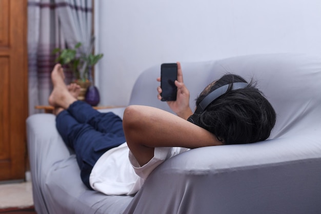 自宅のソファに横たわるスマートフォンとヘッドフォンを持つアジア人男性の全身図オンラインチャット