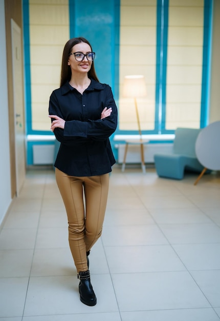 Photo full length view of the attractive woman in dark shirt and glasses standing arms crossed in hall and smiling