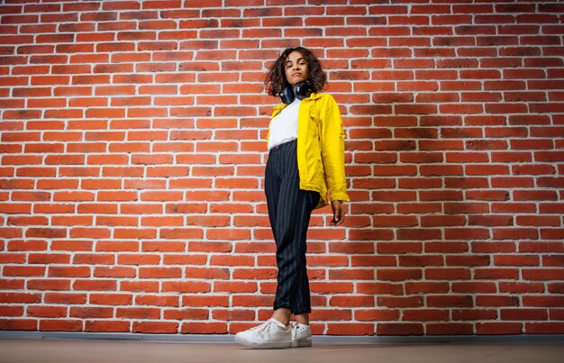 Full length view of the beautiful young sexy girl in a yellow jacket listening to music in headphones on a brick wall background. Excited woman looking at the camera. Positive emotions concept