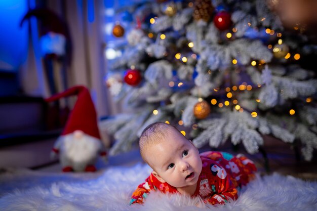 Full length view of the cute baby boy in new year costume lies on the floor Decorative fur tree near the baby Baby looking away