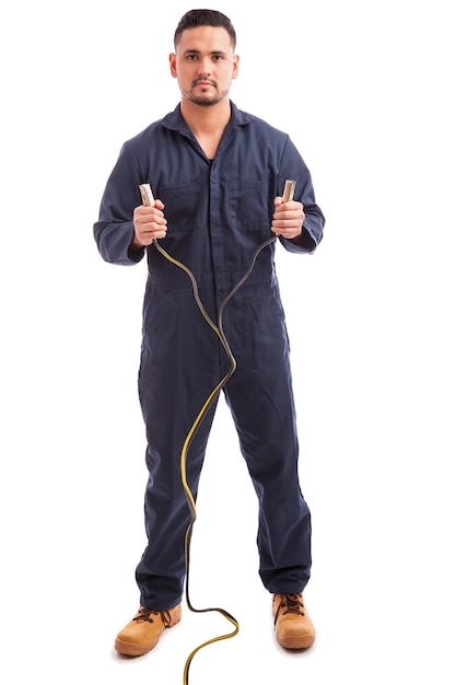 Full length view of a good looking mechanic holding jumper cables and making eye contact against a white background