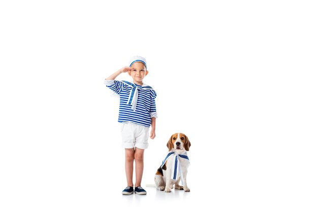 Full length view of preschooler child in sailor costume saluting with smile and beagle dog on white