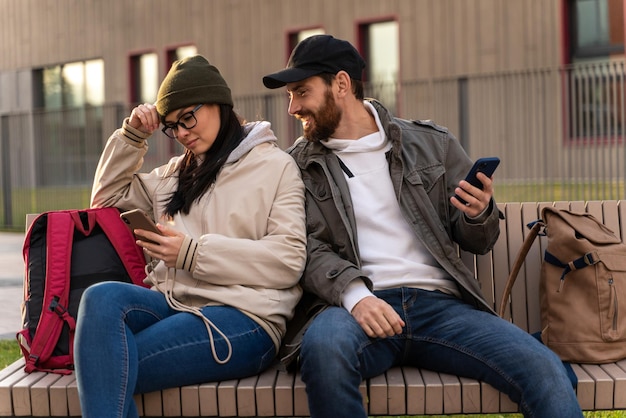 ベンチで通りに一緒に座っている間彼の憤慨している妻のスマートフォンを見ている笑顔の男の全身像