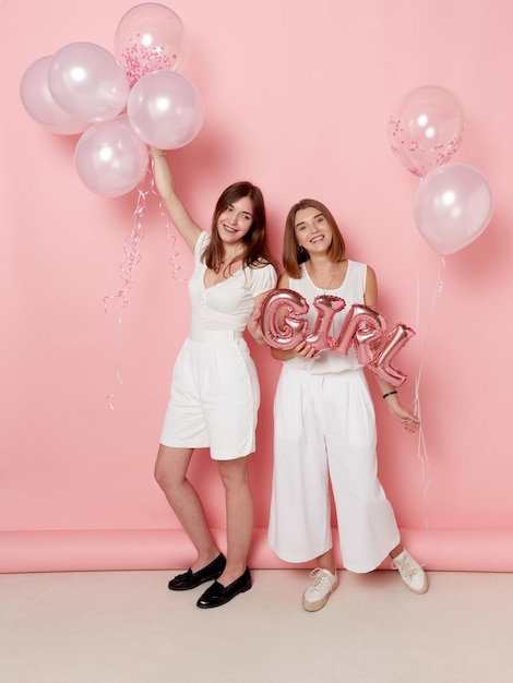 Vista a tutta lunghezza di due ragazze sorridenti vestite di bianco con palloncini su sfondo rosa