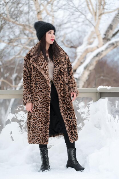 Full length view young female in fur coat with leopard pattern knitted hat black jeans high boots