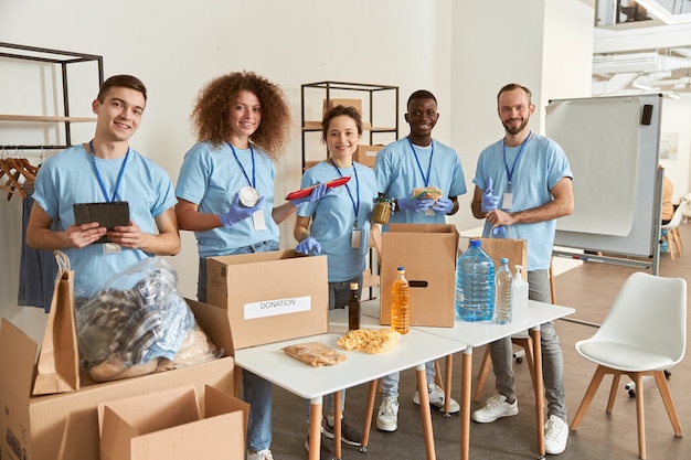 full length of volunteers smiling at camera while sorting packing foodstuff in cardboard boxes