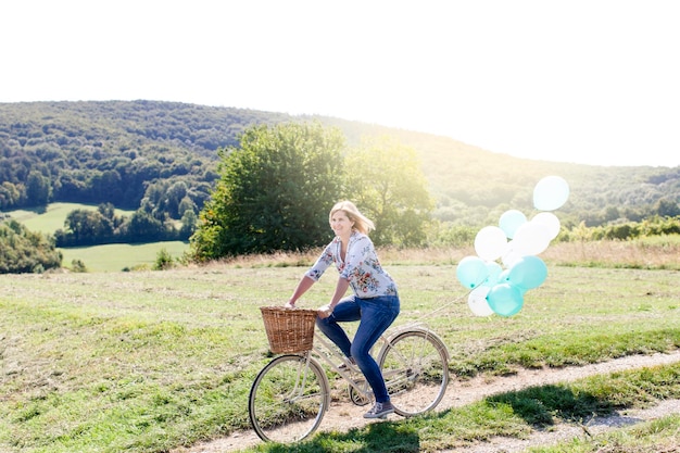 Foto lunghezza completa di una donna in bicicletta con palloncini di elio sul campo durante una giornata di sole