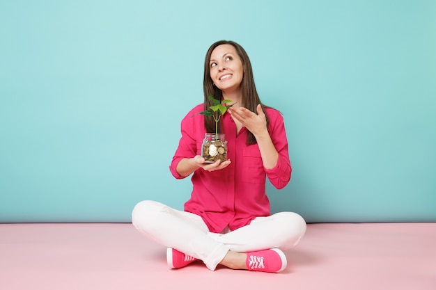 Full length of woman in rose shirt, white pants sitting on floor hold gold coins in glass jar 