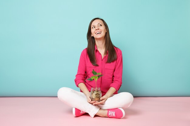 Full length of woman in rose shirt, white pants sitting on floor hold gold coins in glass jar 