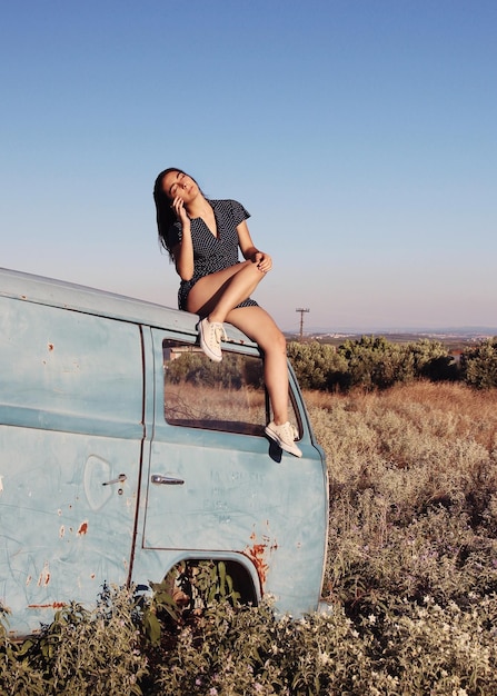 full length of woman sitting on abandoned van at field against clear sky