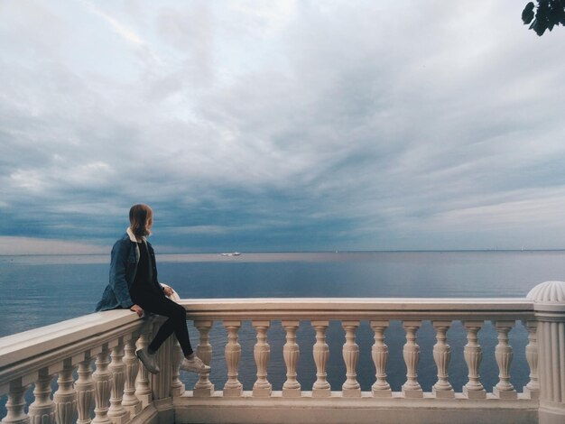 Foto donna a tutta lunghezza seduta su una ringhiera contro il mare.