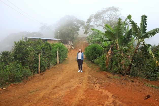 Full length of woman standing on dirt road amidst plants during foggy weather