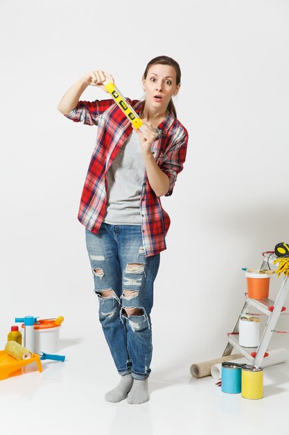 Full length of woman with bubble spirit level standing near instruments for renovation apartment home isolated on white background. Wallpaper, accessories for gluing, painting tools. Repair concept.