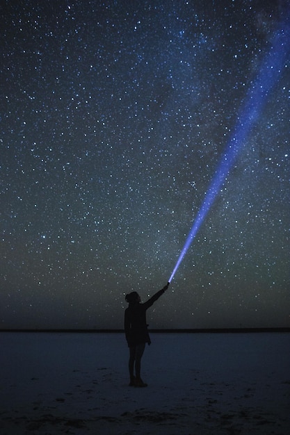 夜に星のフィールドに照らされて立ち向かっている手電灯を持った女性の全長