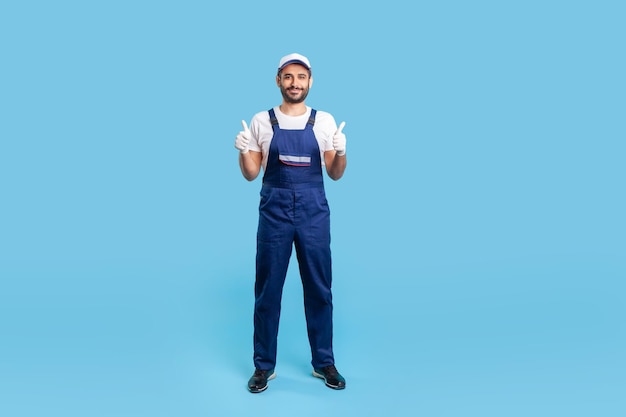 Photo full length workman in overalls cap and protective gloves standing showing thumbs up like gesture and smiling recommending good house repair services studio shot isolated on blue background