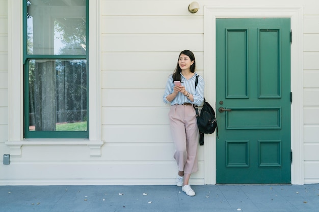 full length young asian korean woman holding smartphone smiling networking by green wooden door and white wall in classic city street outdoors. Female using technology recreation lifestyle copy space