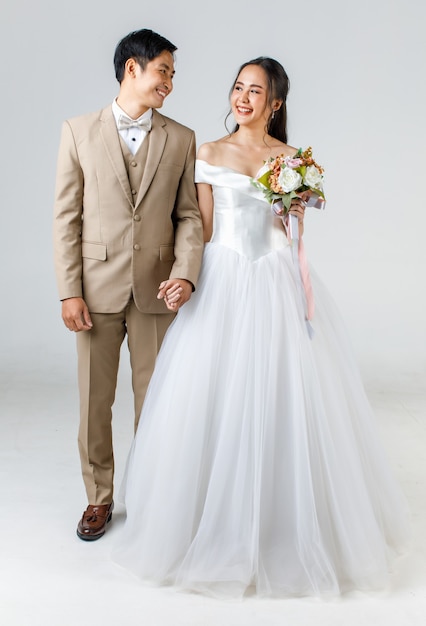 Full length of young attractive Asian couple, man wearing beige suit, woman wearing white wedding gown standing together holding hands. Concept for pre wedding photography.