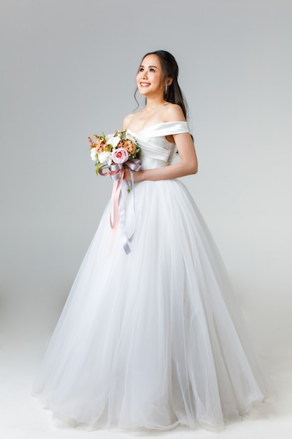 Full length of young attractive Asian woman, soon to be bride, wearing white wedding gown looking happy holding bouquet of flower. Concept for pre wedding photography.