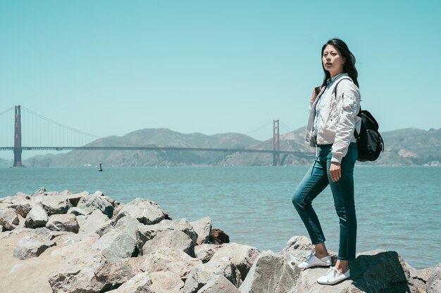 full length of young girl backpacker standing on stone rocks near the sea on the bay sightseeing under sunshine in summer america. red golden state bridge in background with nature amazing mountain.