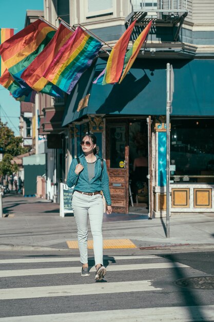 full length of young girl lesbian walking on sunny day on zebra cross in modern city castro area. corner cafe shop hanging on rainbow flags and welcome same sex lover customer. woman on crosswalk