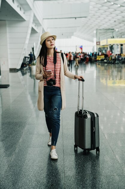 Full length of young girl traveler in hat walking with hand in\
cell phone and luggage suitcase in airport lobby. asian elegant\
lady in casual wear with camera searching direction of departure\
gate.