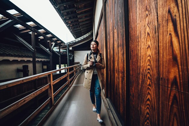 full length young girl traveler recording tour trip in kyoto japan by slr camera. chinese lady walking in corridor in japanese old building along the wooden wall indoor. asian woman sightseeing.