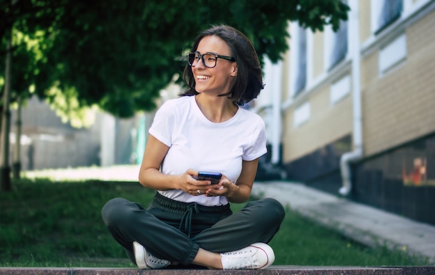 Full-length  of a young girl, with the glasses on her face, holding a phone in her hands, smiling, enjoying herself outside.