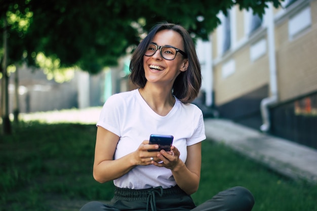 眼鏡をかけ、携帯電話を手に持ち、笑顔で外で楽しんでいる少女の全身像。