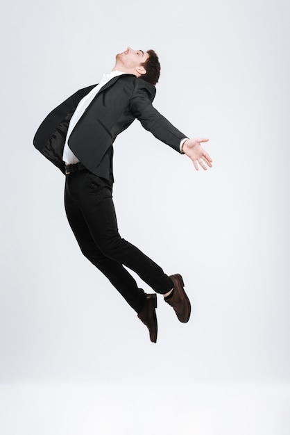 Full length young happy business man in black suit flying  isolated on a white wall