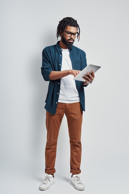 Молодой человек в очках с помощью цифрового планшета, стоя у серой стены в полный рост