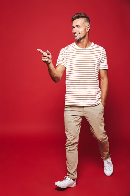 Full length of young man in striped t-shirt gesturing index fingers aside at copyspace isolated on red