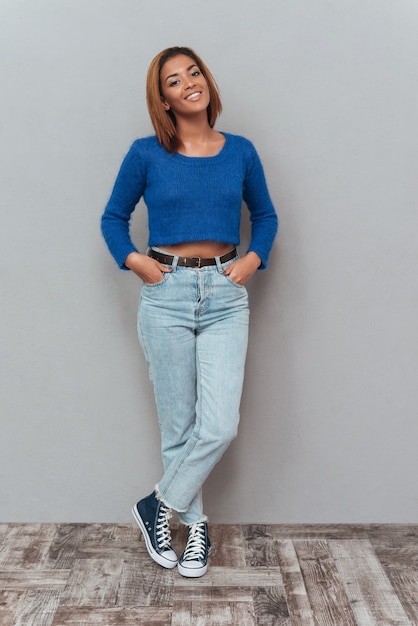 Foto giovane donna africana sorridente integrale in maglione e jeans che tiene le braccia in tasca e in piedi vicino al muro.