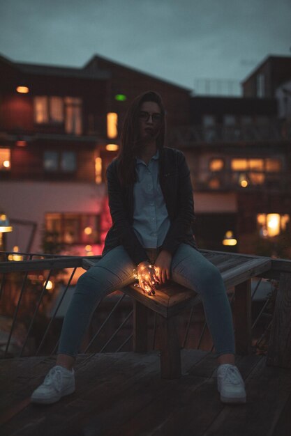 Photo full length of young woman holding illuminated lights while sitting against building at night
