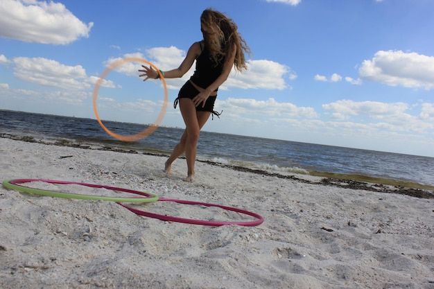 Foto lunghezza completa di una giovane donna che gioca con un cerchio di plastica sulla spiaggia contro un cielo nuvoloso