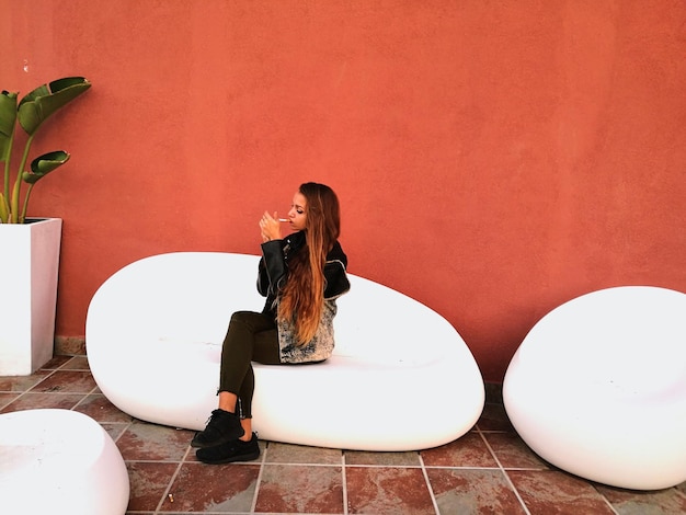 Photo full length of young woman smoking cigarette against wall on seat