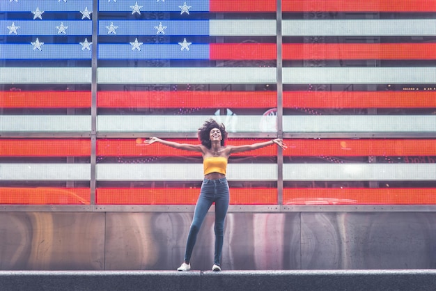 Photo full length of young woman standing with arms outstretched against neon american flag in city