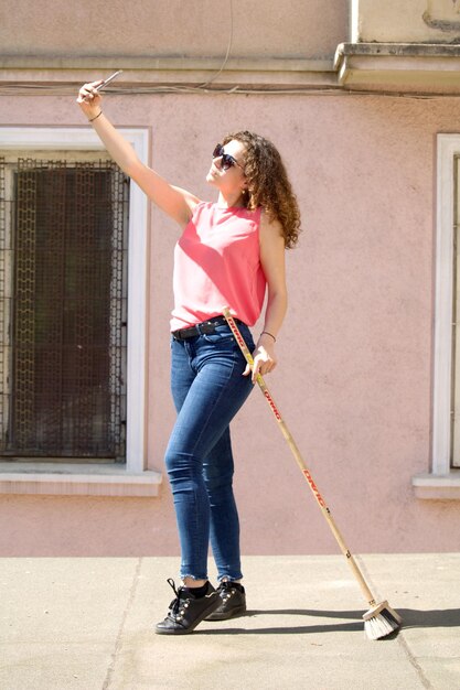 Full length of young woman taking selfie while holding broom on street in city during sunny day