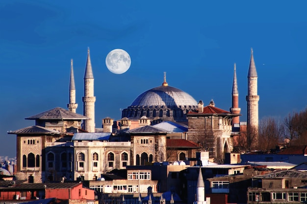 A full moon rises over a city with a large blue mosque in the foreground.