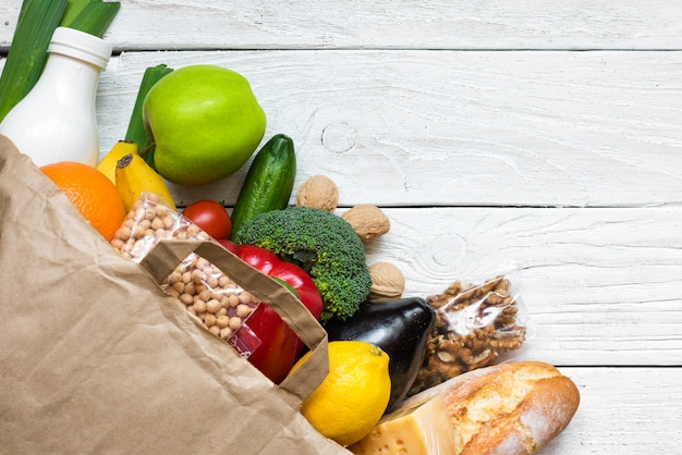 Full paper bag of different healthy vegetarian food on white wooden background. fruits, vegetables, nuts, bread and milk