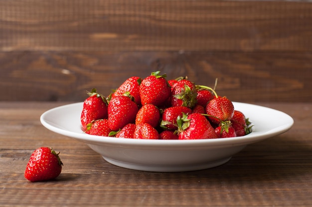 Full plate of ripe fresh strawberries and one strawberry on the brown wooden table