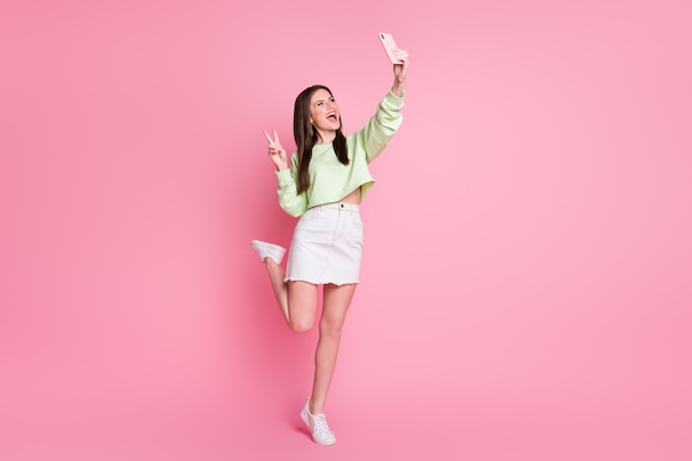 Full size photo of charming lady hold telephone excited blogging make selfies show v-sign symbol wear casual green crop pullover jeans short skirt shoes isolated pink pastel color background
