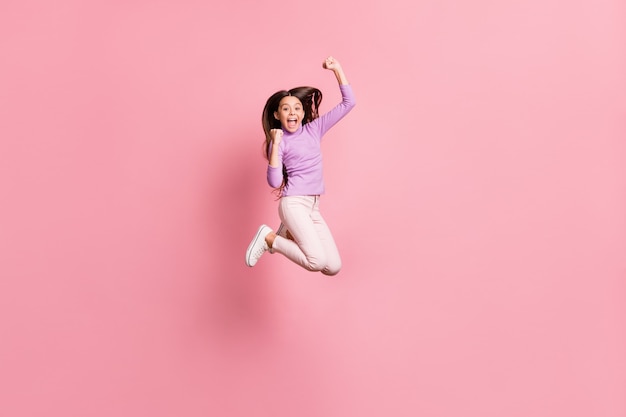 Full size photo of ecstatic little girl jump raise fists wear purple sweater isolated over pastel color background