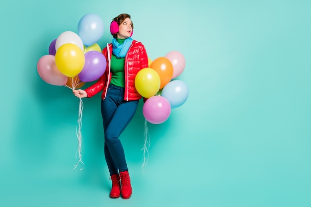 Full size photo of funny lady hold many colorful air balloons\
students party look empty space wear casual red coat scarf pink ear\
covers pants shoes isolated teal color wall