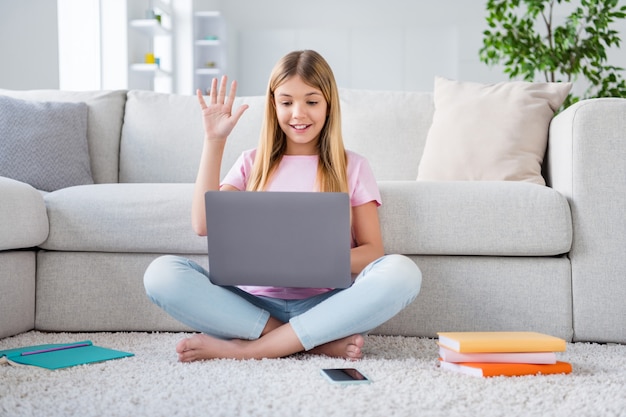 Full size photo of positive kid girl sit floor legs crossed study remote have online laptop tutor education conference meeting greet in house indoors