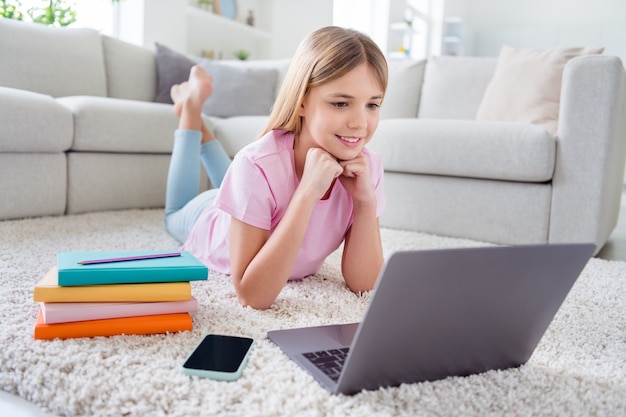 Full size photo of positive little kid girl study remote laptop\
watch tutor education webinar lying floor carpet in house\
indoors